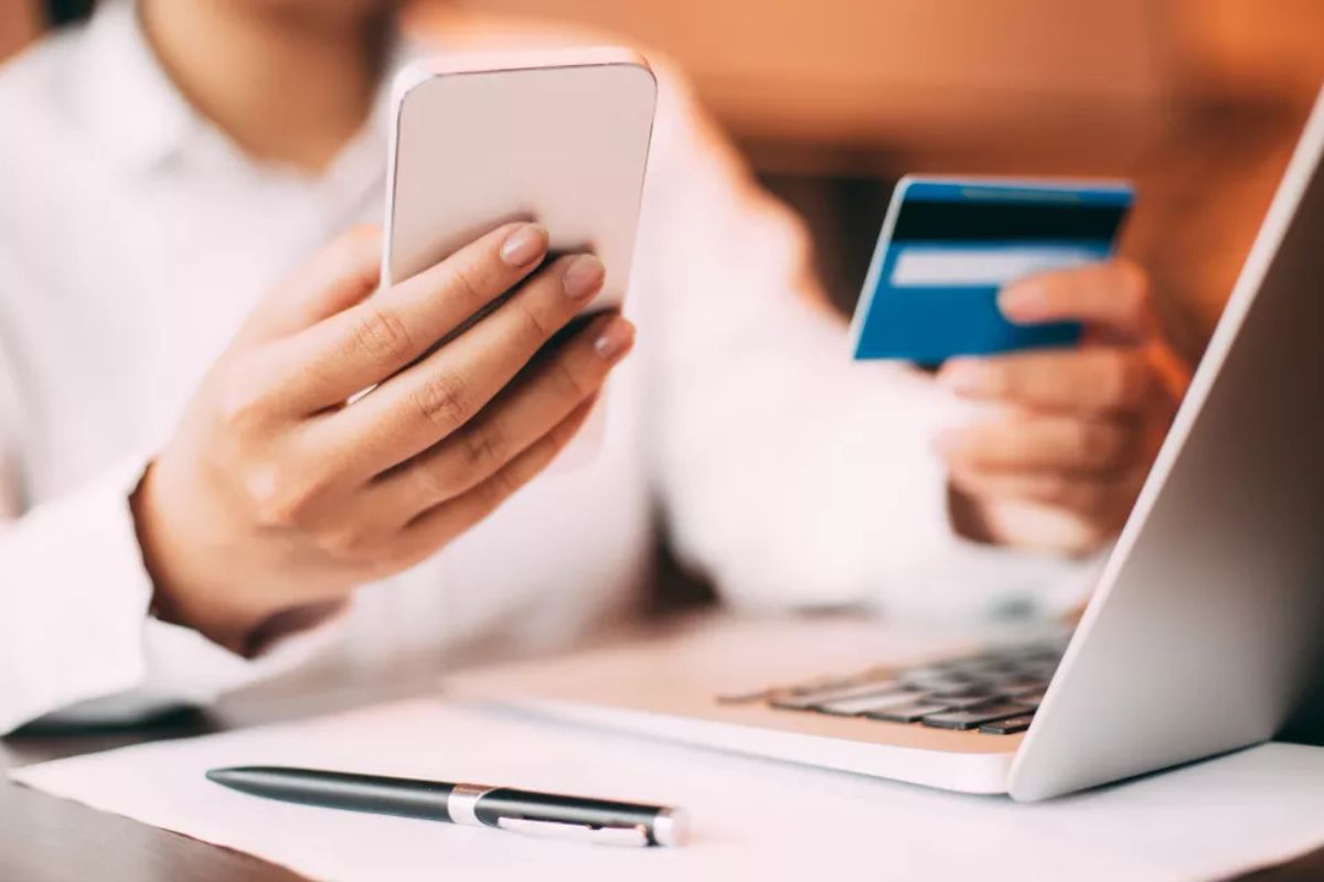 Consumidor mexendo no celular e com a mão no cartão de crédito em frente ao notebook, fazendo compra online.