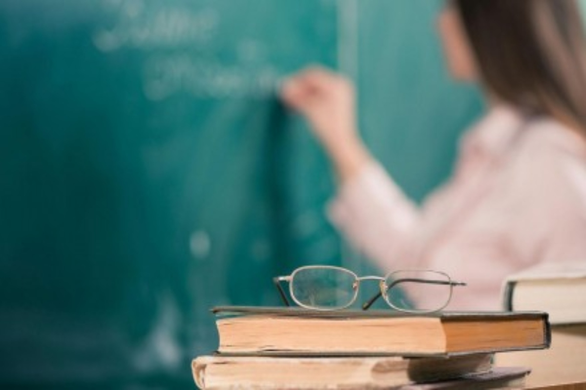 Livros, óculos e professora escrevendo no quadro; processo seletivo para professores está aberto (Foto: Reprodução/Google)